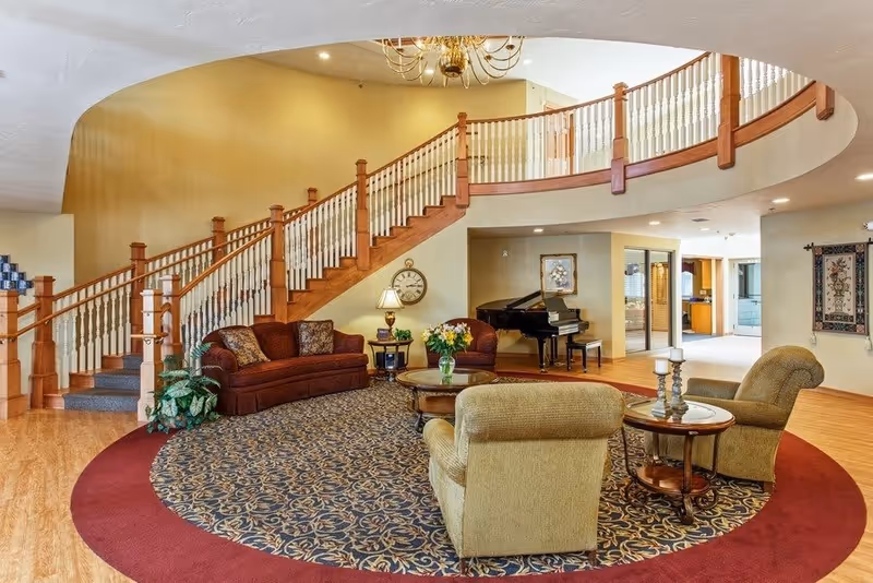 A spacious living room area with a large curved staircase featuring wooden railings and white balusters. The room has a patterned carpet with a red border, a brown sofa, two beige armchairs, a round wooden coffee table with a vase of flowers, and a side table with a lamp. A black grand piano is placed against the far wall, and a chandelier hangs from the ceiling.