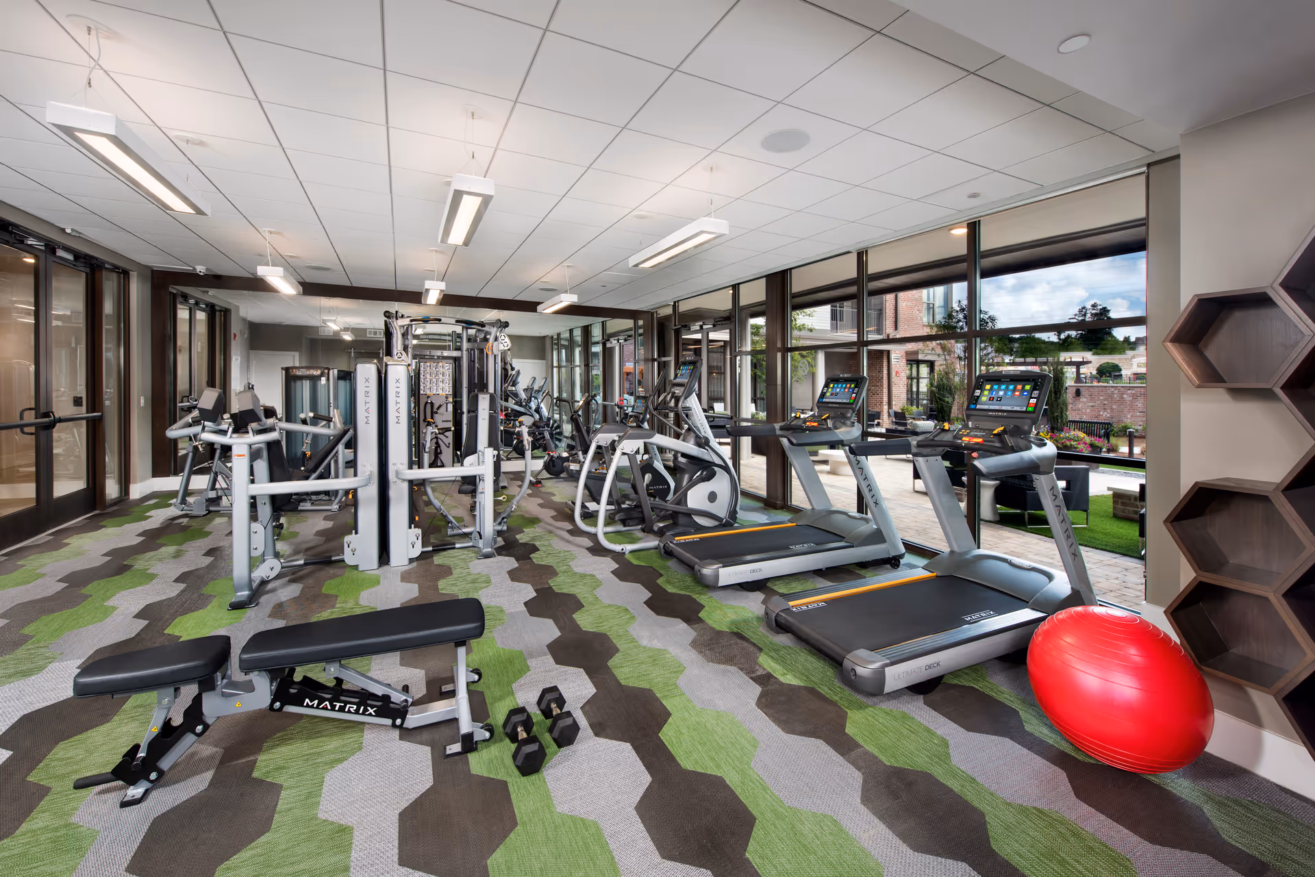A modern fitness room with various exercise equipment including treadmills, elliptical machines, a weight bench, dumbbells, and a multi-functional gym machine. The room has large windows offering a view of an outdoor patio area with seating and greenery. The floor is covered with a patterned carpet in shades of green, gray, and brown. A red exercise ball is placed near hexagonal wooden shelves on the right wall.