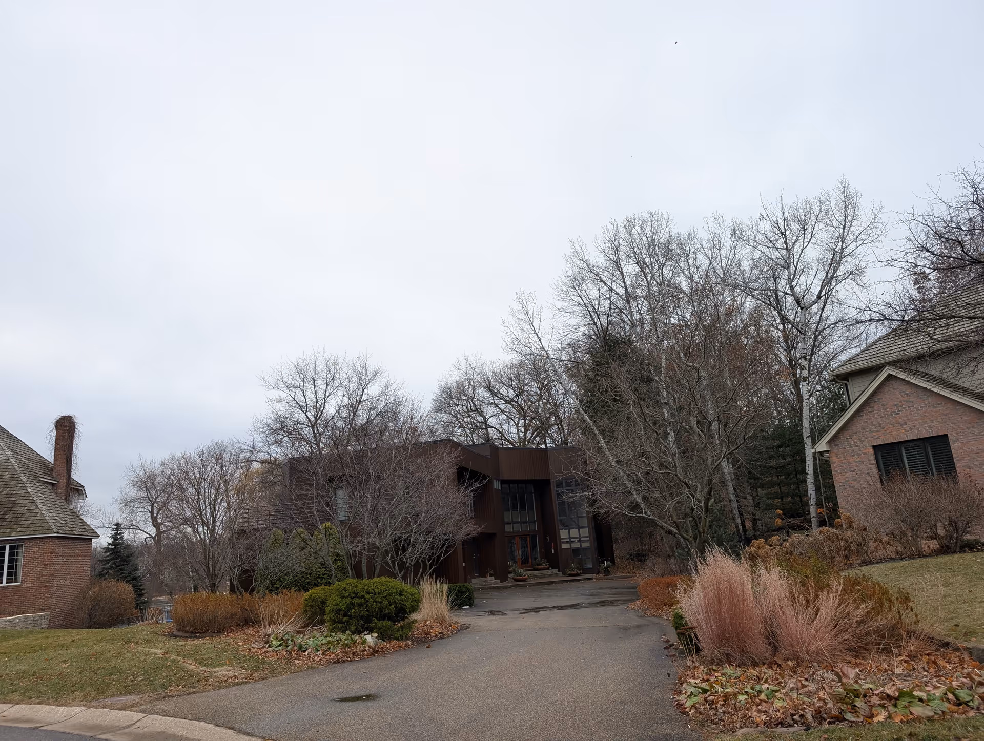 Front exterior of a brick and dark-paneled residential building with a driveway, winter landscaping, and bare trees under an overcast sky.