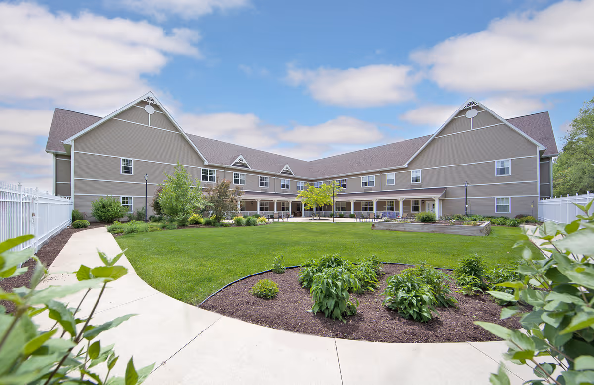 A large, two-story senior living facility building with beige siding and white trim surrounds a well-maintained green lawn and garden area. There is a curved concrete walkway leading through the garden with various shrubs and plants. The sky is partly cloudy with blue patches visible.