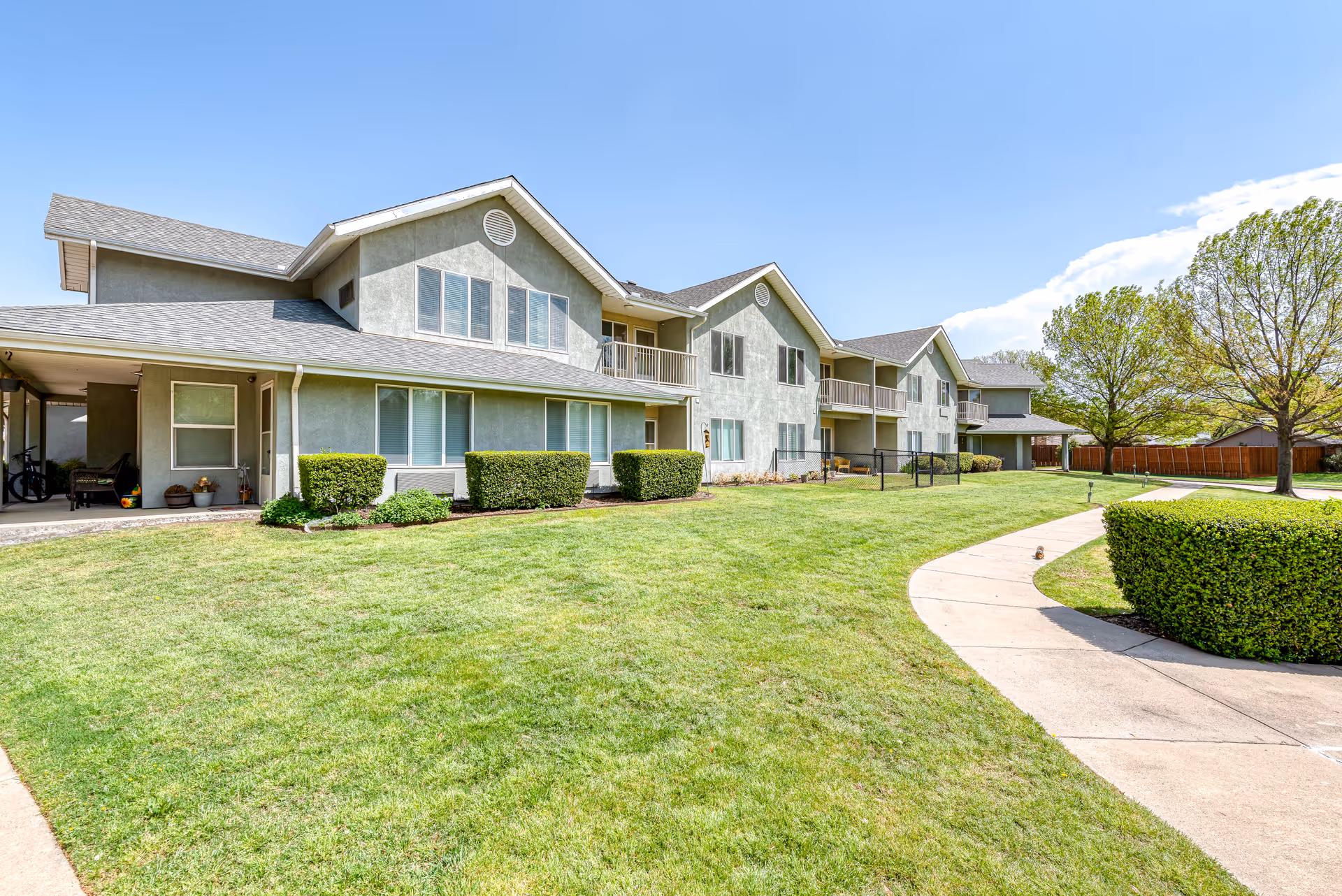 Exterior view of a two-story senior living facility building with gray walls and multiple windows. The building is surrounded by a well-maintained green lawn, trimmed bushes, a curved concrete walkway, and several trees under a clear blue sky.