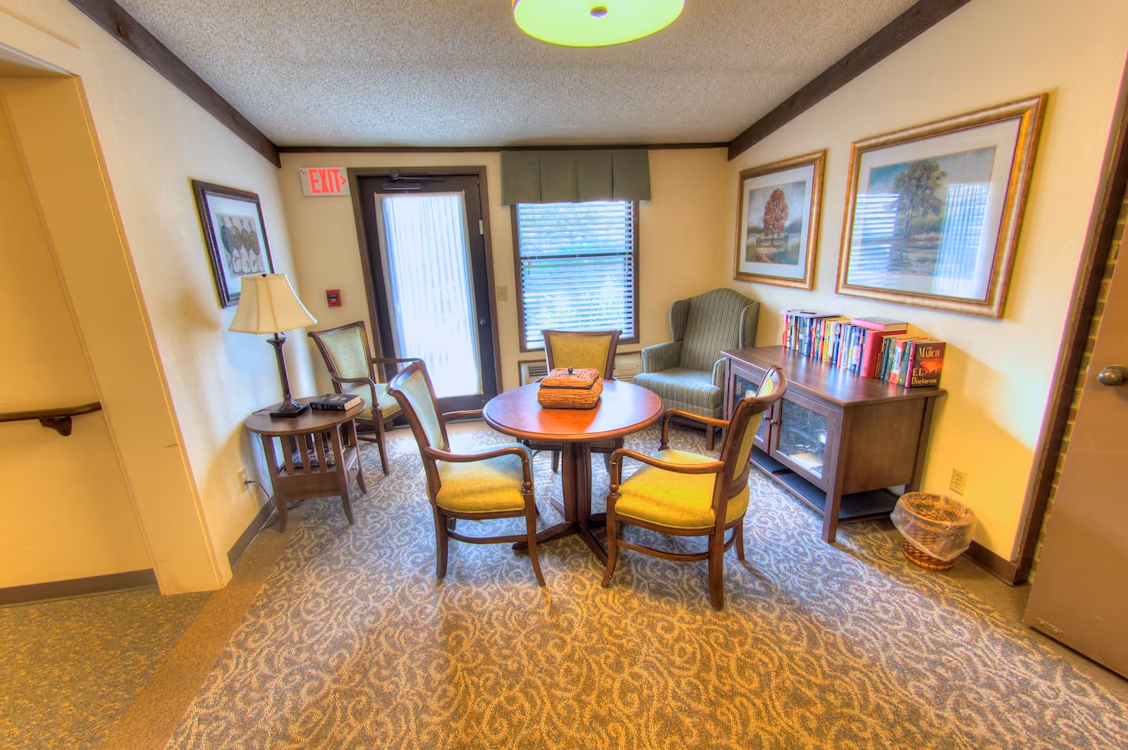 A cozy sitting area in Springfield Assisted Living featuring a round wooden table with four cushioned chairs around it. There is a green upholstered armchair near a window with blinds, a wooden side table with a lamp and a phone, and a wooden cabinet with books on top. The walls have framed artwork, and the floor is carpeted with a patterned design. A door with a window and an exit sign is visible in the background.