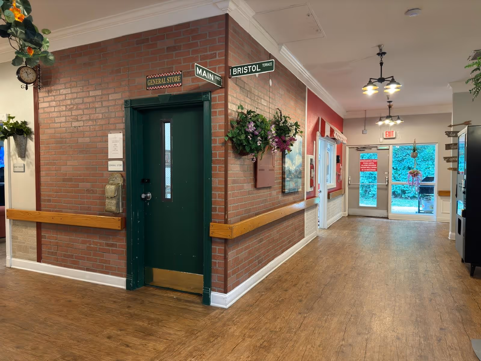 Interior hallway of a senior living facility with wood flooring and brick walls. A green door labeled 'General Store' is on the left corner with street signs above it reading 'Main Street' and 'Bristol Terrace.' There are hanging flower baskets on the walls, a clock, and an exit door at the end of the hallway with a window showing greenery outside.
