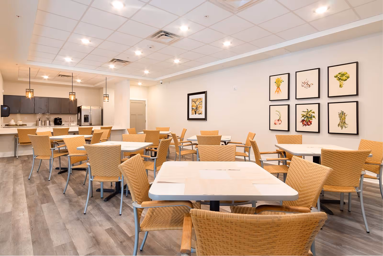 A bright dining room with multiple square tables surrounded by wicker chairs. The room features a modern kitchen area with dark cabinets, a stainless steel refrigerator, and pendant lights hanging over a counter with bar stools. The walls are decorated with framed botanical prints of vegetables and plants. The floor has a wood-like finish and the ceiling has recessed lighting.