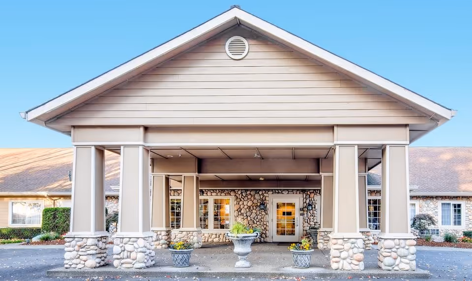 Front exterior view of Cedar Crest Alzheimer's Special Care Center featuring a covered entrance with stone pillars and a peaked roof, potted plants, and windows on either side of the main door under a clear blue sky.