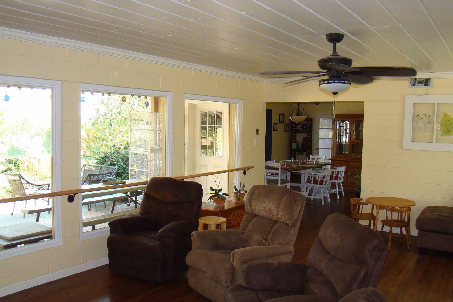 A cozy living room area with three brown recliner chairs facing a large window that looks out onto a patio with outdoor seating. The room has light yellow walls, a ceiling fan with lights, wooden floors, and a small wooden table with chairs. In the background, there is a dining area with a white table and chairs, and a wooden cabinet.