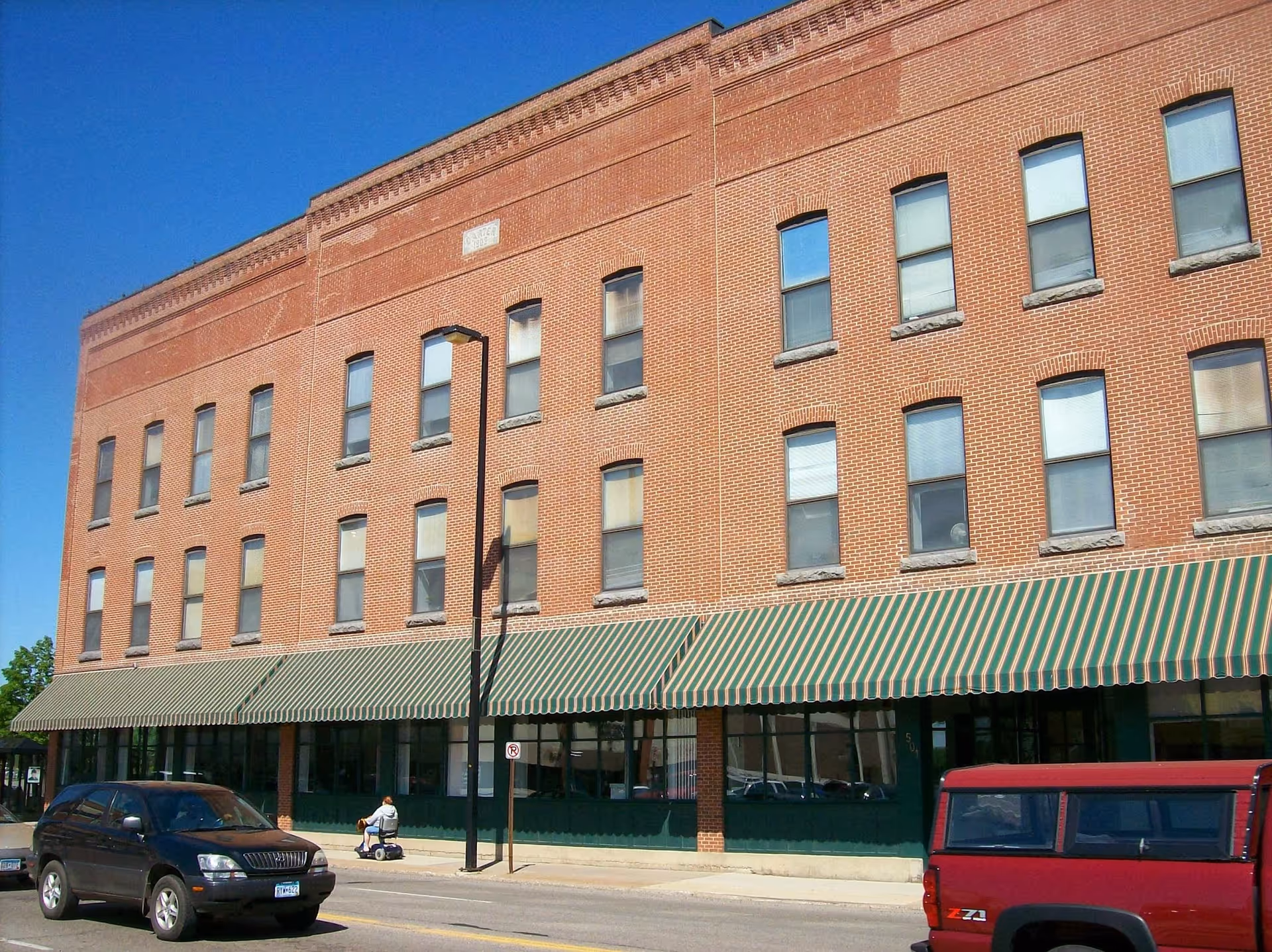 Exterior view of a three-story brick building with multiple windows and green and white striped awnings over the ground floor windows. Two vehicles are parked on the street in front of the building, and a person on a mobility scooter is on the sidewalk. The sky is clear and blue.