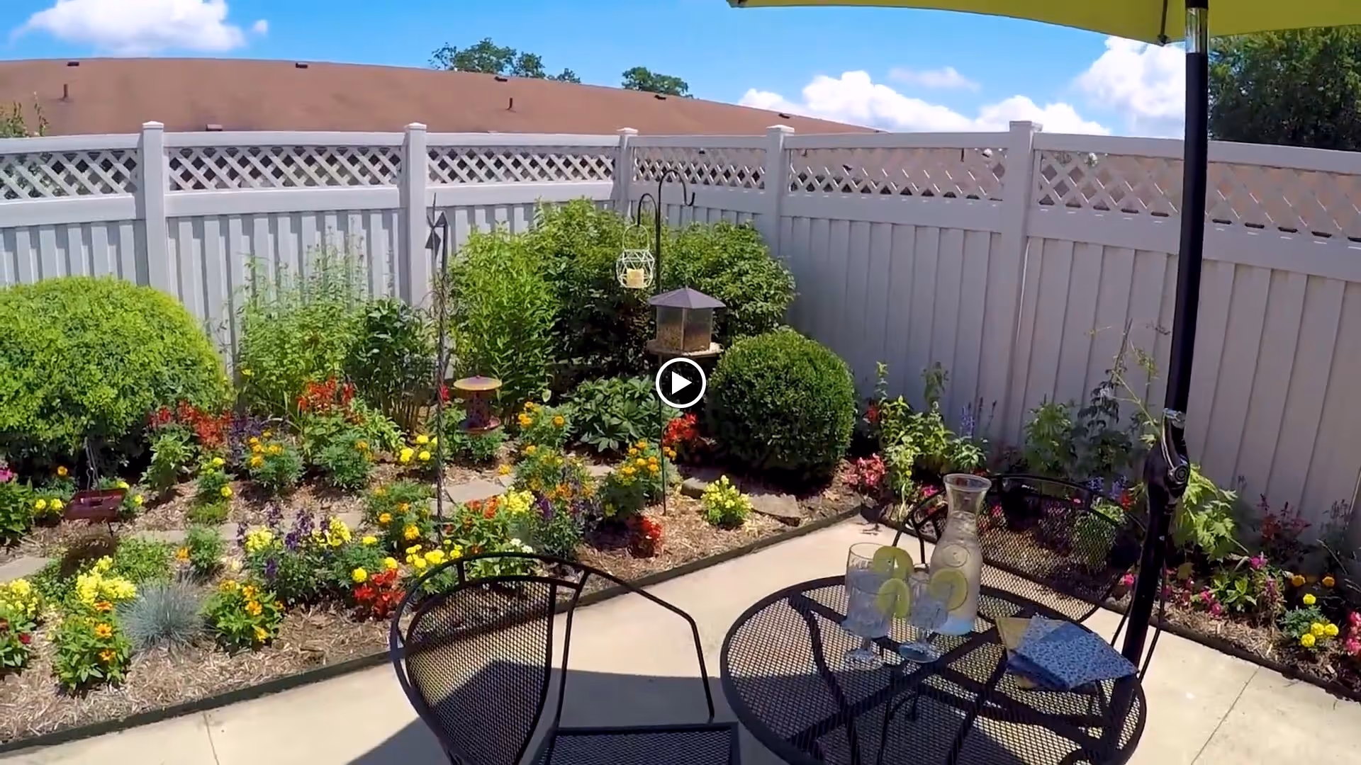 A sunny fenced patio garden with a metal table and chairs under an umbrella, a pitcher with lemon slices, and colorful flowerbeds along a white fence.