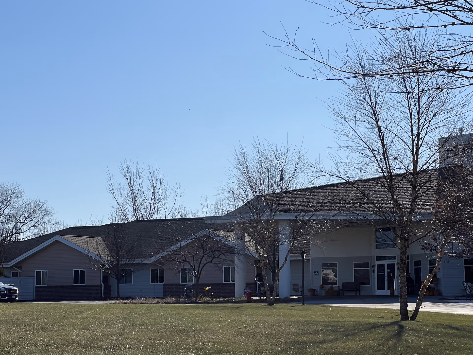 Exterior view of Milan Estates senior living facility on a clear day, showing a single-story building with a covered entrance, leafless trees, and a grassy lawn in the foreground.