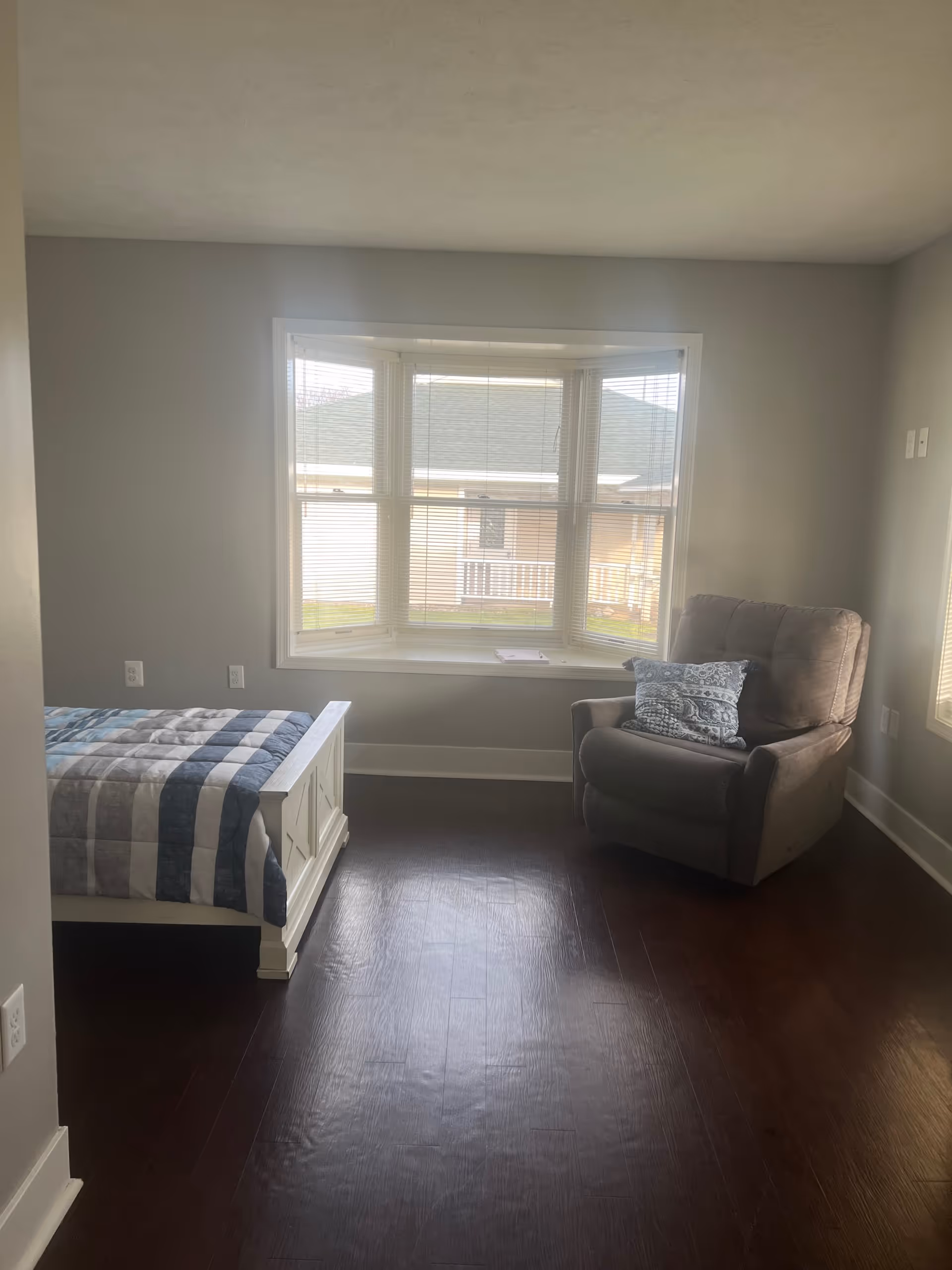 A bedroom with a large window featuring blinds, a white bed frame with a blue and gray striped quilt, and a gray armchair with a patterned pillow. The room has dark wood flooring and light gray walls.