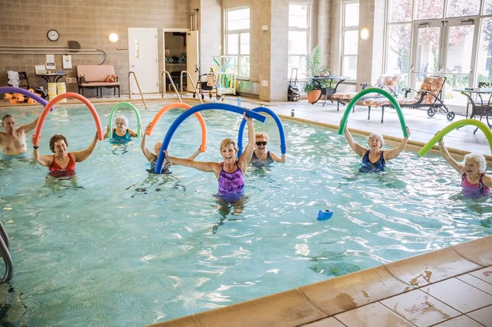 A group of elderly people participating in a water exercise class in an indoor swimming pool, each holding colorful pool noodles above their heads. The pool area is well-lit with large windows and has seating and plants around the perimeter.