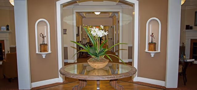 Interior view of a senior living facility hallway with a round wooden table in the foreground holding a potted plant with white flowers. The hallway is framed by two alcoves each containing a small statue. The walls are painted beige with white trim, and there is seating visible in the background.