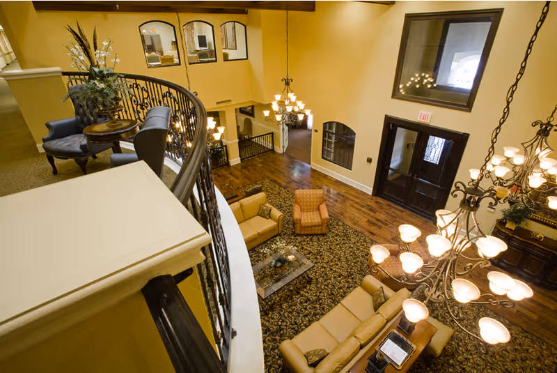 View from an upper level overlooking a spacious living room area with beige sofas, an armchair, a patterned carpet, and a glass coffee table. The room features warm yellow walls, large windows, elegant chandeliers, and a decorative wrought iron railing along the balcony.
