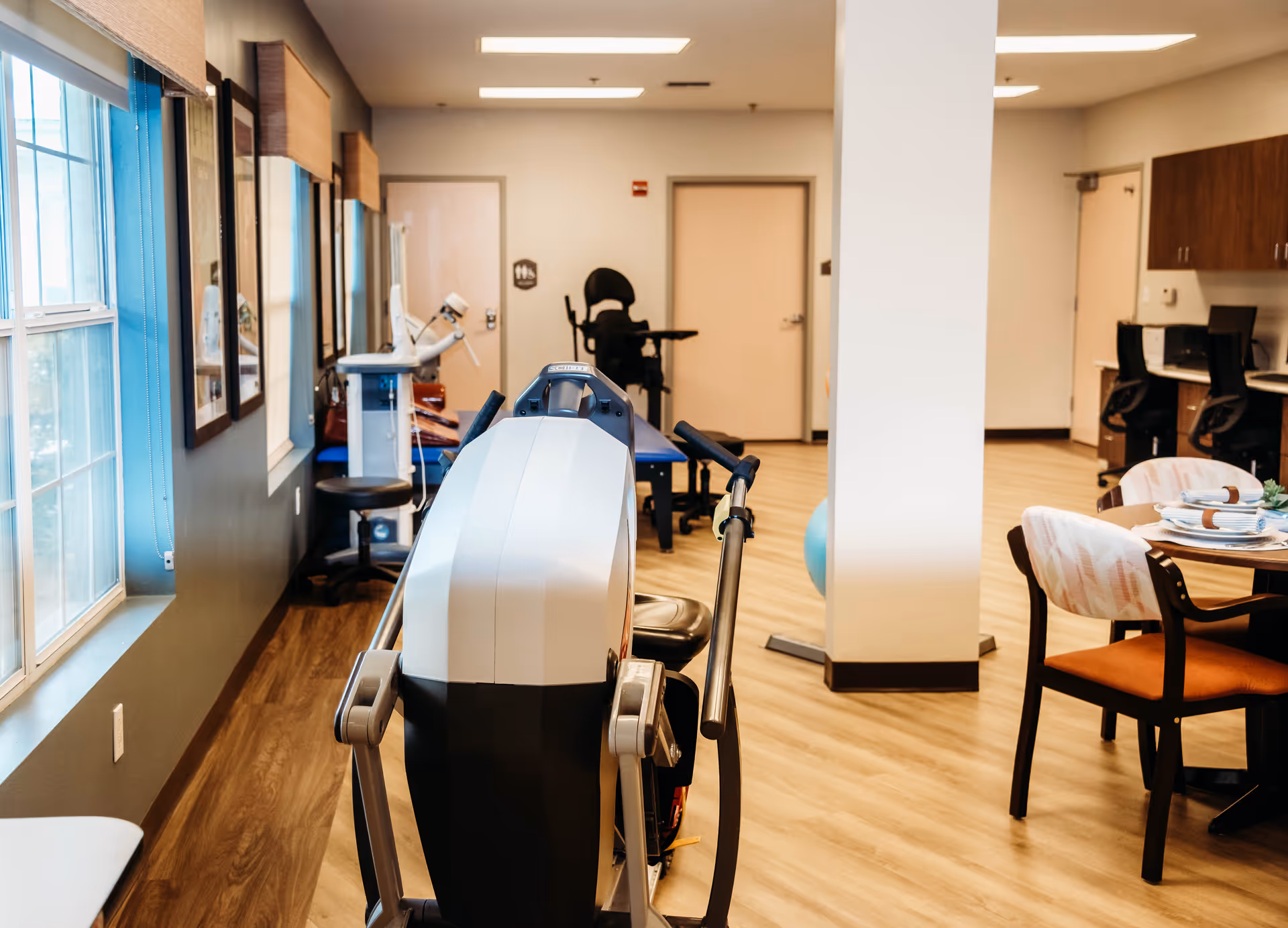 Rehabilitation/common room with exercise equipment, a dining table and chairs, and windows along one wall.