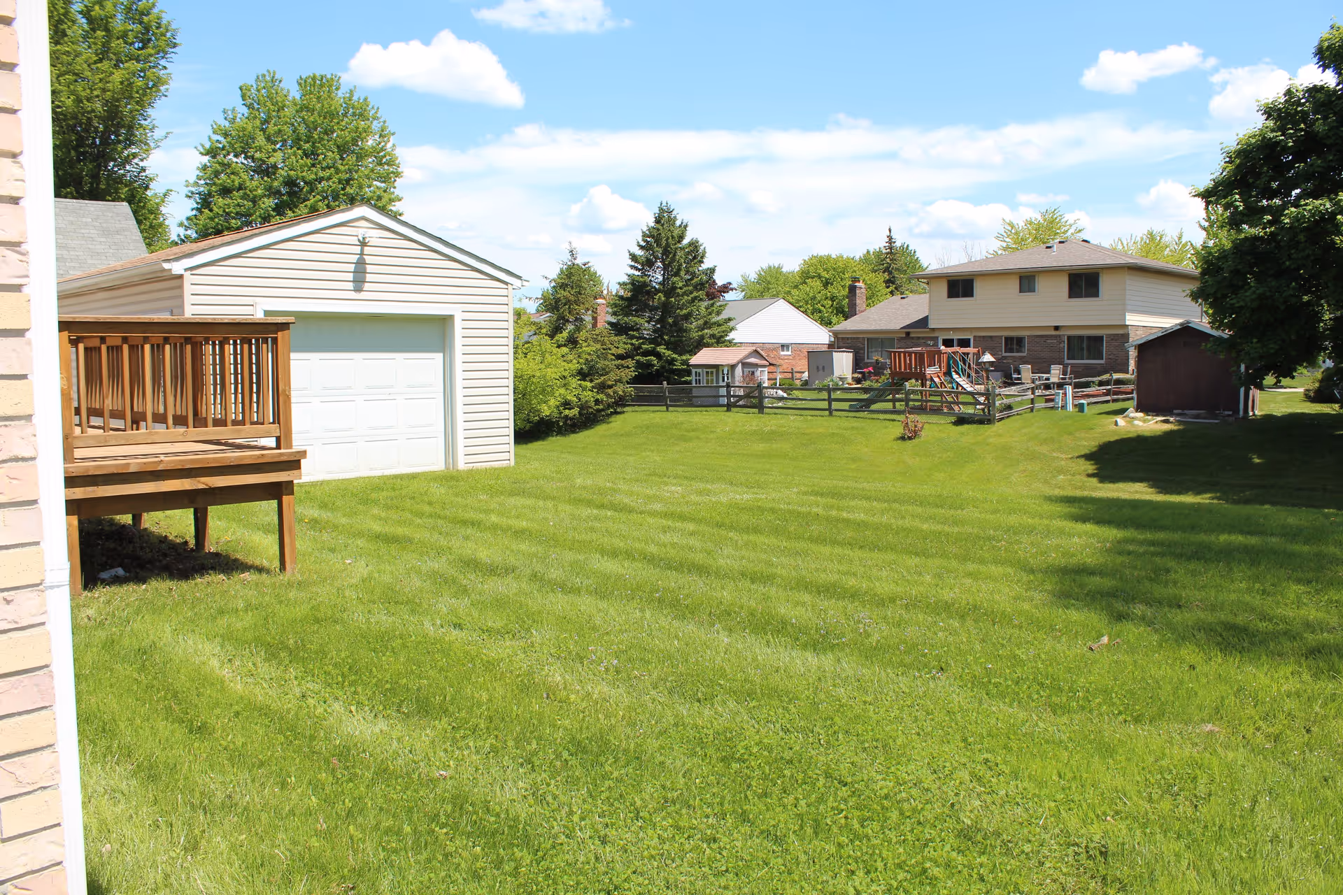 A spacious green backyard with a wooden deck attached to a building on the left, a white garage with a closed door in the middle, and houses with fenced yards and playground equipment in the background under a partly cloudy blue sky.