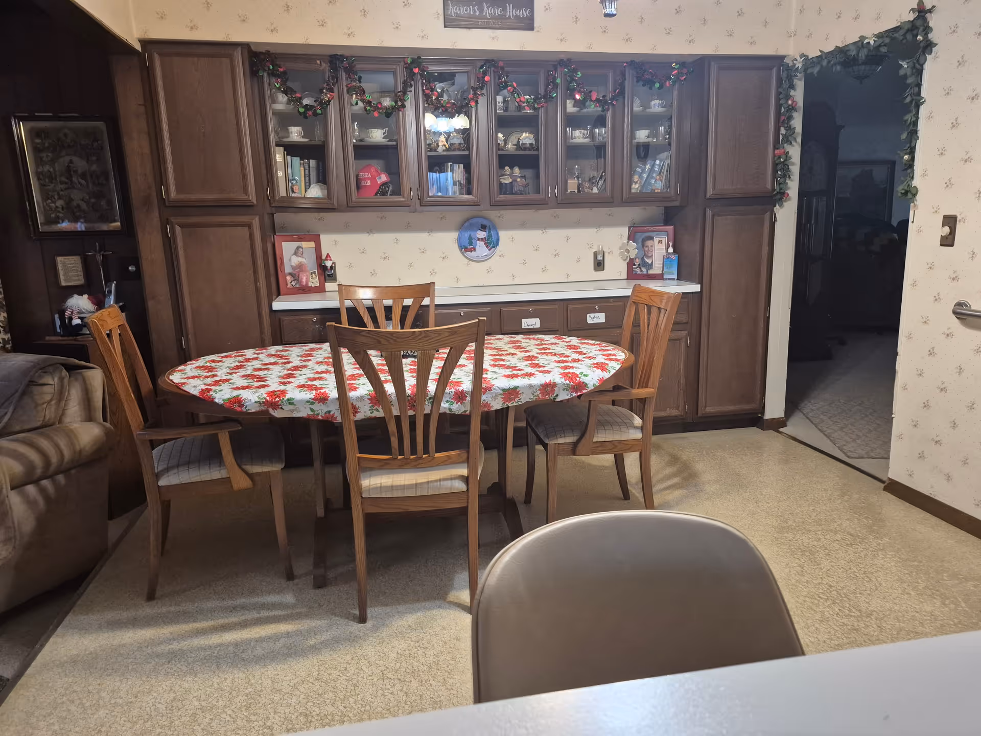 A dining area with a wooden oval table covered with a red and white floral tablecloth surrounded by four wooden chairs with cushioned seats. Behind the table is a wall with dark wooden cabinets and glass-fronted shelves displaying various decorative items and framed photos. The room has beige wallpaper with a subtle pattern and a garland decoration above the cabinets. To the left, part of a beige sofa is visible, and to the right, there is an open doorway leading to another dimly lit room.