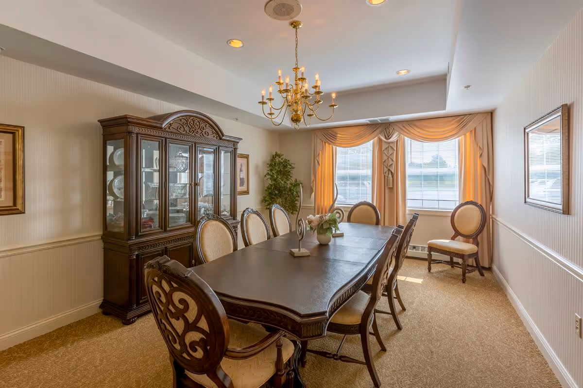 A formal dining room with a long dark wooden table surrounded by eight upholstered chairs. A large wooden china cabinet with glass doors displaying dishes is against the left wall. The room has beige carpet, cream-colored walls with wainscoting, and a window with beige curtains letting in natural light. A gold chandelier hangs above the table, and framed artwork decorates the walls.