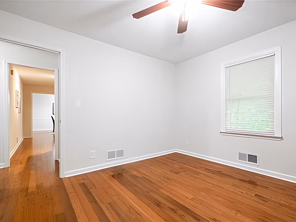 Empty room with hardwood floors, a ceiling fan, a window with blinds, and a doorway leading to a hallway.