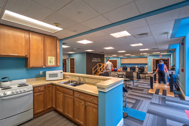 Open interior common area with a kitchenette in the foreground and a rehabilitation/fitness space with equipment and people in the background.