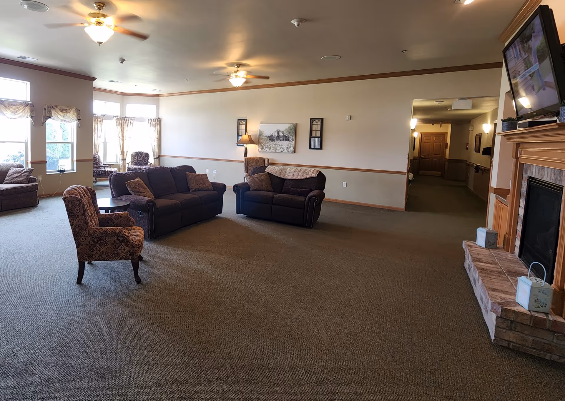 A spacious living room area in a senior living facility with multiple upholstered sofas and armchairs arranged around a central space. The room has large windows with curtains allowing natural light, ceiling fans with lights, a fireplace with a mounted TV above it, and a hallway leading to other rooms.