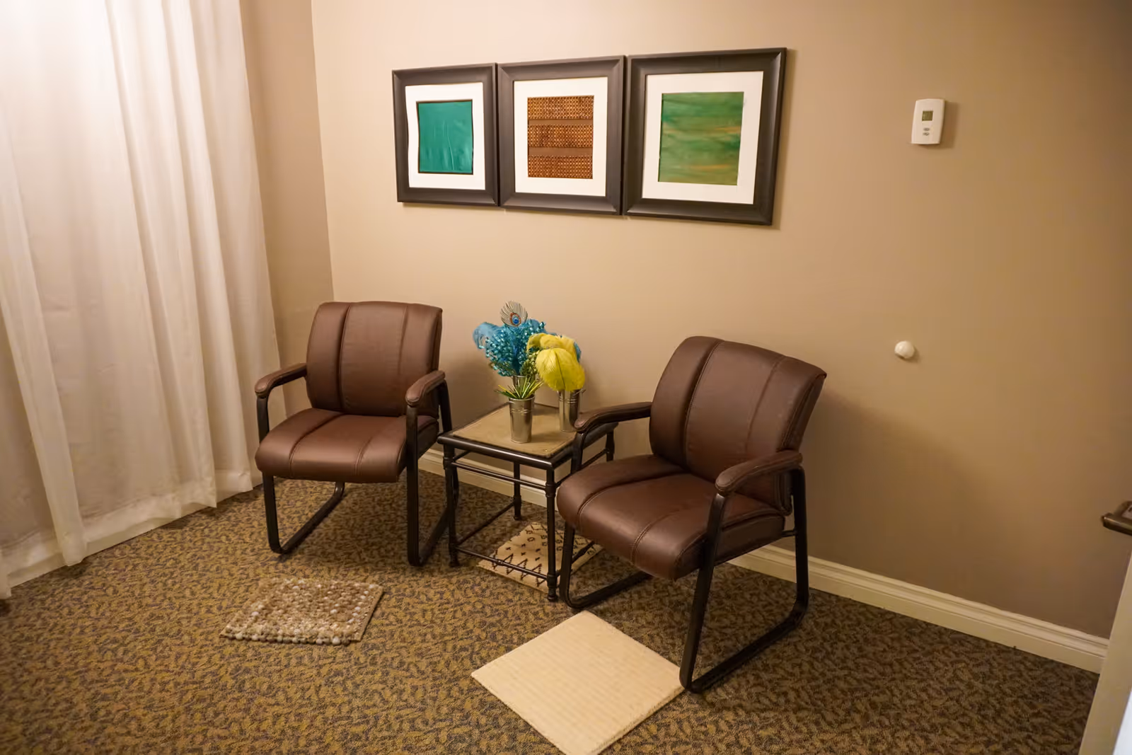 A cozy seating area with two brown cushioned chairs placed on a patterned carpet. Between the chairs is a small glass-top side table holding two small flower arrangements with blue and yellow flowers. Behind the chairs on the beige wall are three framed abstract art pieces in green and brown tones. A white curtain hangs to the left side of the image.
