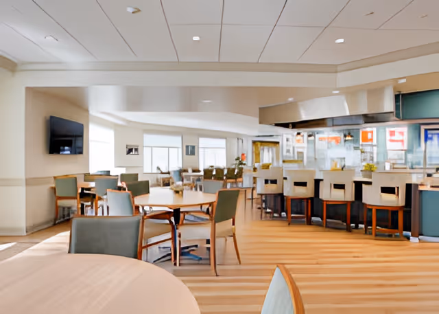 A spacious dining area in a senior living facility with multiple round tables and chairs arranged neatly. There is a bar counter with high chairs on the right side and large windows in the background allowing natural light to fill the room. The floor has a striped pattern and the ceiling has recessed lighting.