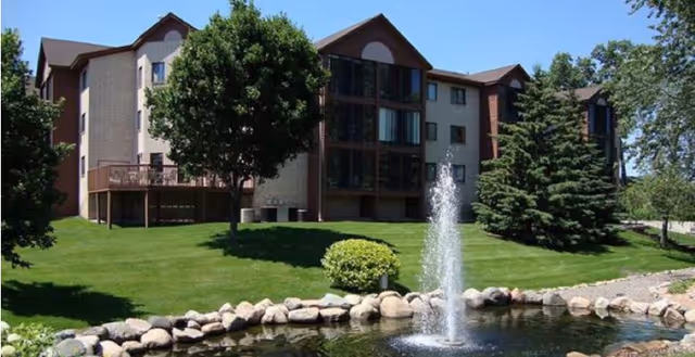 Exterior view of a multi-story senior living facility building with beige and brown walls, surrounded by green grass, trees, and a small pond with a water fountain in the foreground under a clear blue sky.