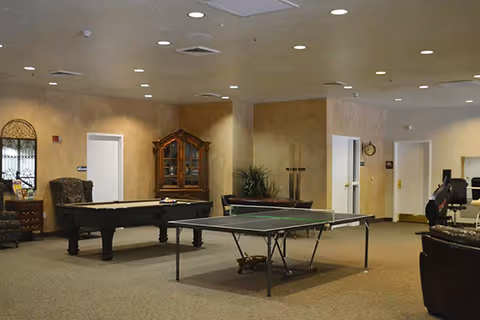 A spacious recreational room with a pool table and a ping pong table in the center. The room has beige walls and carpeted floors. There is a wooden cabinet against the wall, a few chairs, and some exercise equipment visible on the right side. The ceiling has recessed lighting.