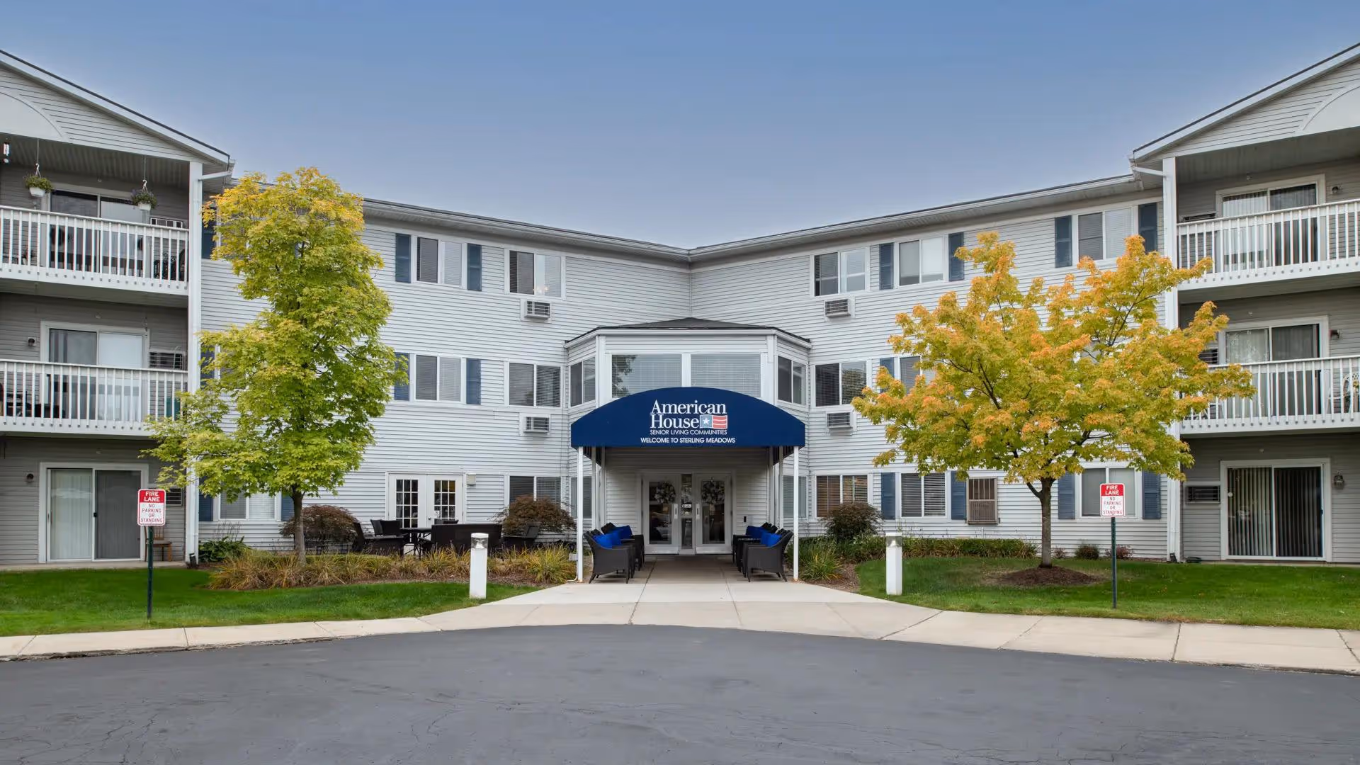 Front entrance of a three-story senior living building with a blue 'American House' canopy, balconies on either side, and trees in the lawn.