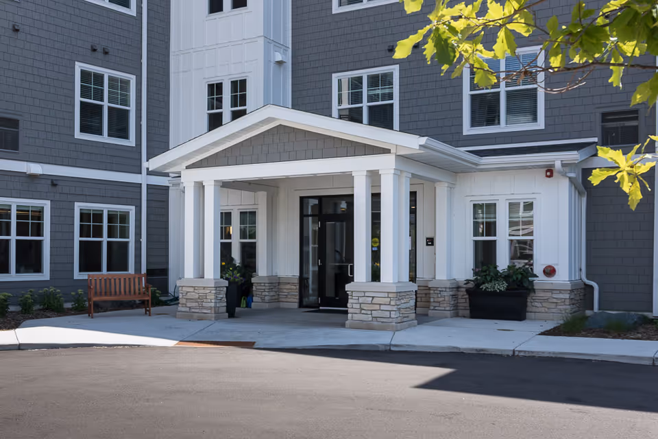 Entrance of a senior living facility with a covered porch supported by white columns and stone bases. The building exterior is gray with white trim around the windows. There is a wooden bench to the left and potted plants near the entrance. A tree branch with green leaves is visible in the upper right corner.