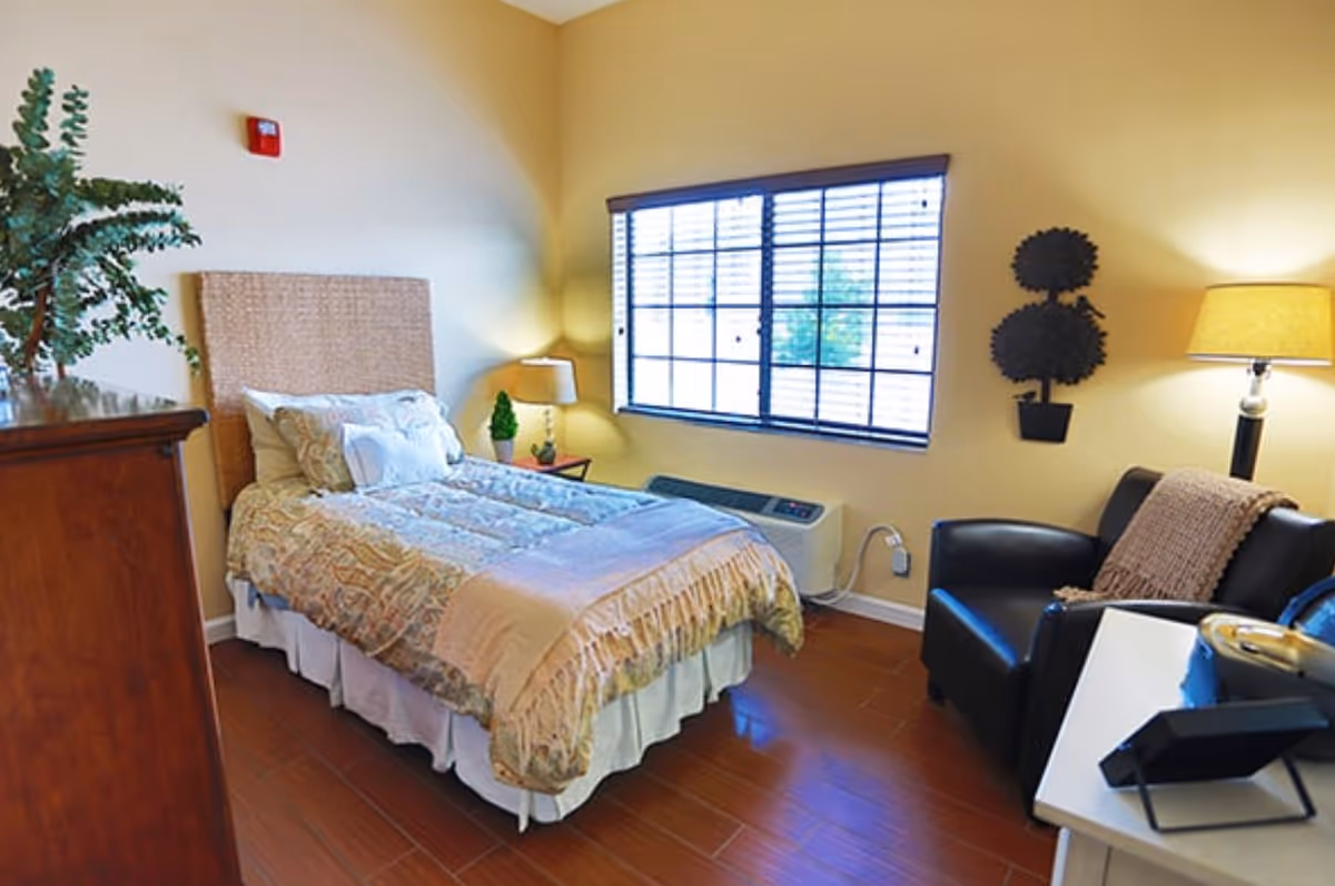 A cozy bedroom with a single bed covered in a patterned comforter and pillows. There is a wooden dresser with a plant on top, a window with blinds letting in natural light, a small side table with a lamp and a small plant, a black armchair with a throw blanket, and a floor lamp providing warm lighting.
