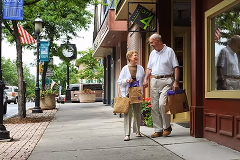 An elderly couple walking on a sidewalk in a small town shopping area, carrying shopping bags and smiling at each other. Trees, street lamps, and parked cars line the street.