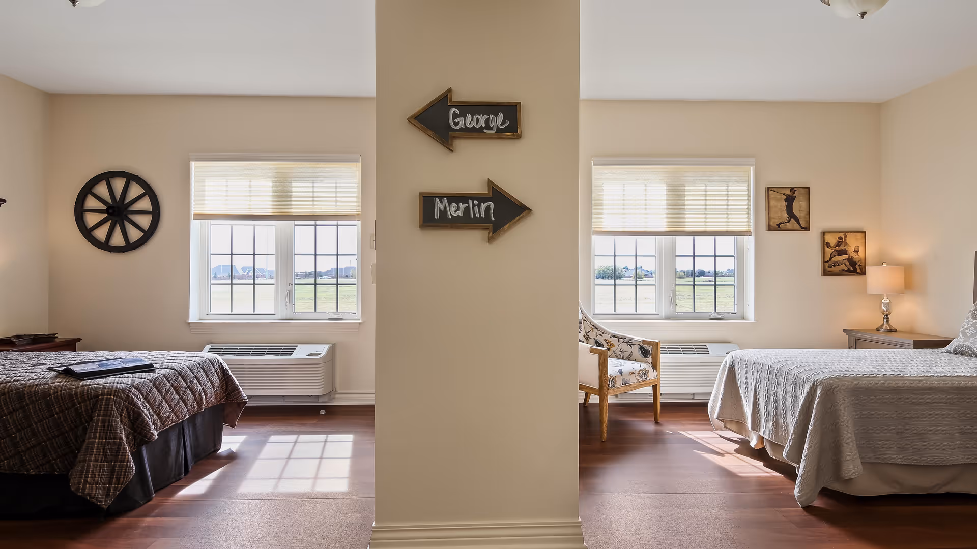A shared bedroom in a senior living facility with two separate beds on either side of a central wall. Each side has a window with blinds, a heating/cooling unit below the window, and decorative wall art. The left side features a dark plaid bedspread and a circular wall decoration, while the right side has a light-colored bedspread, a wooden chair with floral upholstery, a bedside table with a lamp, and two framed pictures on the wall. Two small arrow signs on the central wall point to each side, labeled 'George' and 'Merlin'.