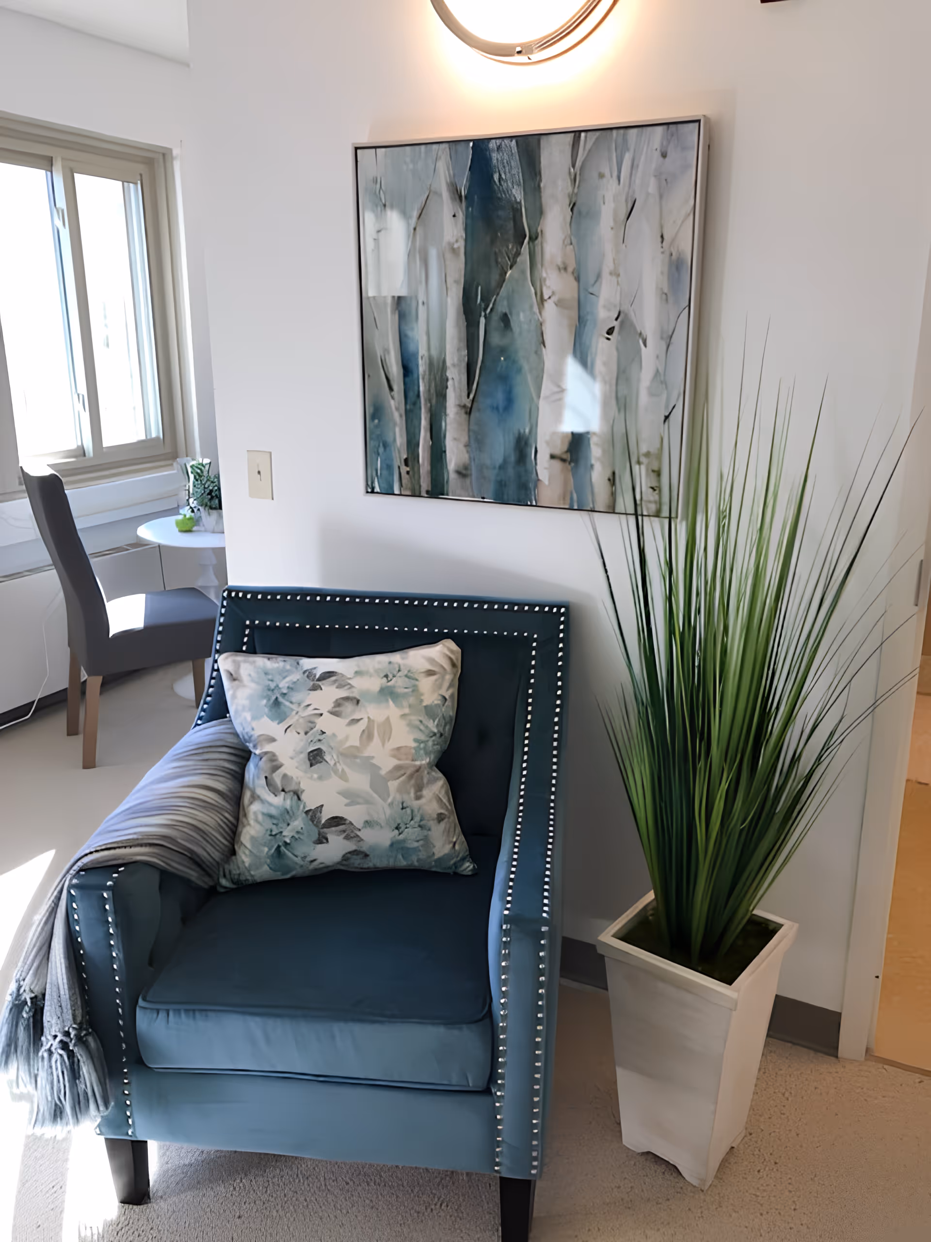 A blue studded armchair with a floral pillow and throw next to a tall potted plant and abstract wall art by a window.