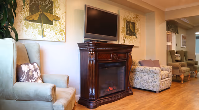 A cozy living room area with a wooden electric fireplace and a flat-screen TV mounted above it. The room features comfortable armchairs with floral and solid cushions, wooden flooring, and decorative wall art with a dragonfly theme. The space is warmly lit and has a welcoming atmosphere.