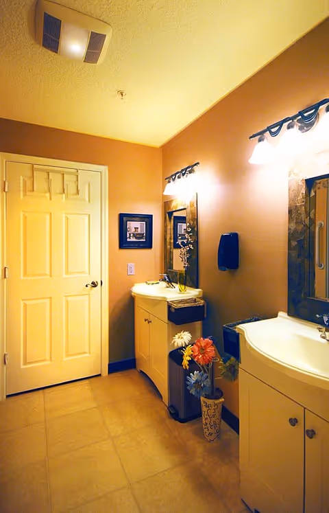 Well-lit bathroom with two vanities and mirrors, a closed door, and decorative flowers on the floor.