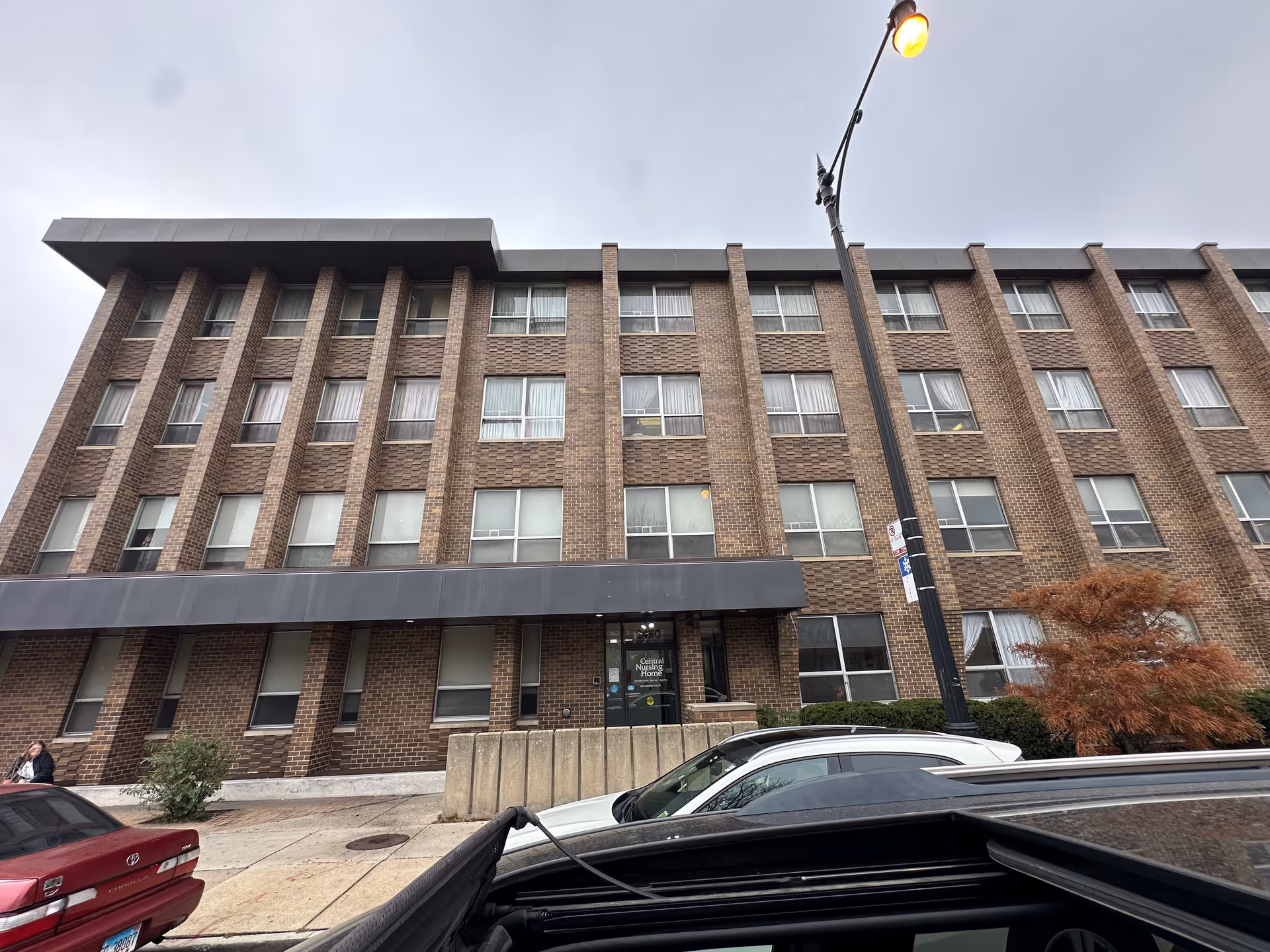 Exterior front view of a multi-story brick building with many windows, a black awning above the entrance, and a street lamp in front. Several cars are parked along the street, and a person is walking on the sidewalk. The entrance door has a sign that reads 'Central Nursing Home'.
