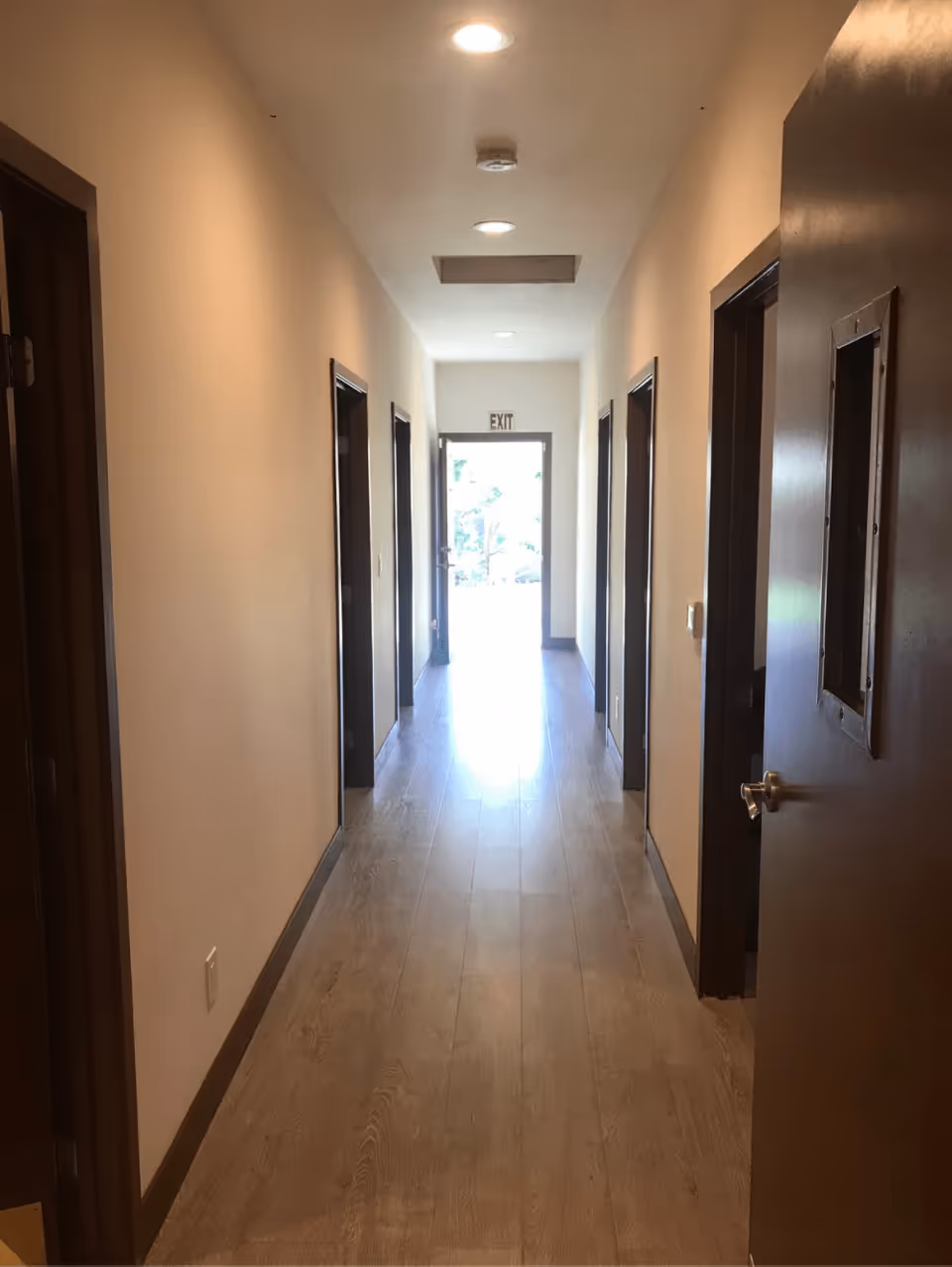 A long, well-lit interior hallway with wooden floors, doors on both sides and an exit door at the far end.
