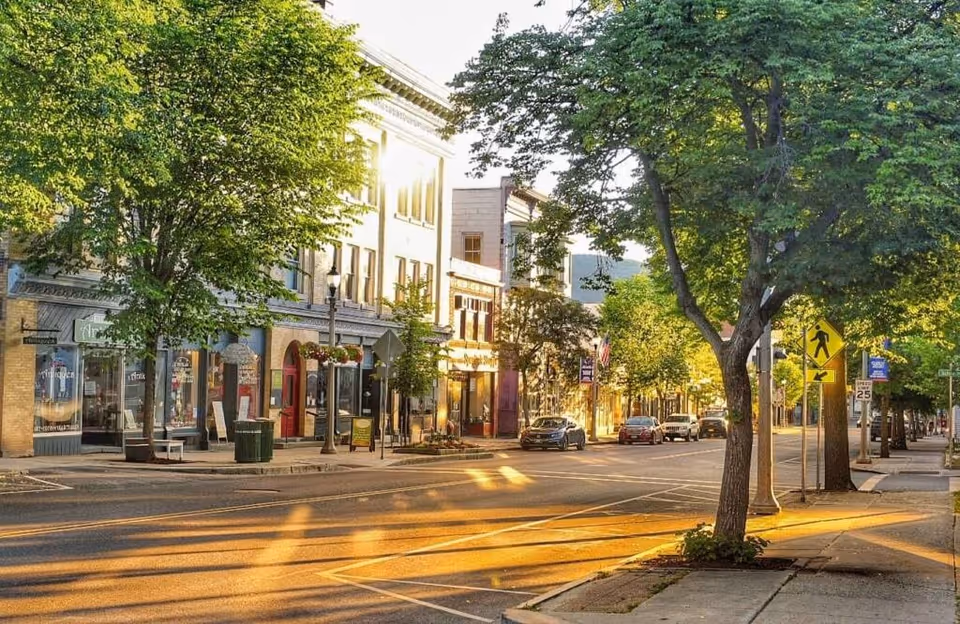 A sunlit street scene in a small town with trees lining the sidewalk and several parked cars. Buildings with storefronts and large windows are visible on the left side of the street, with a pedestrian crossing sign and speed limit sign on the right. The sunlight casts long shadows across the road and sidewalk.
