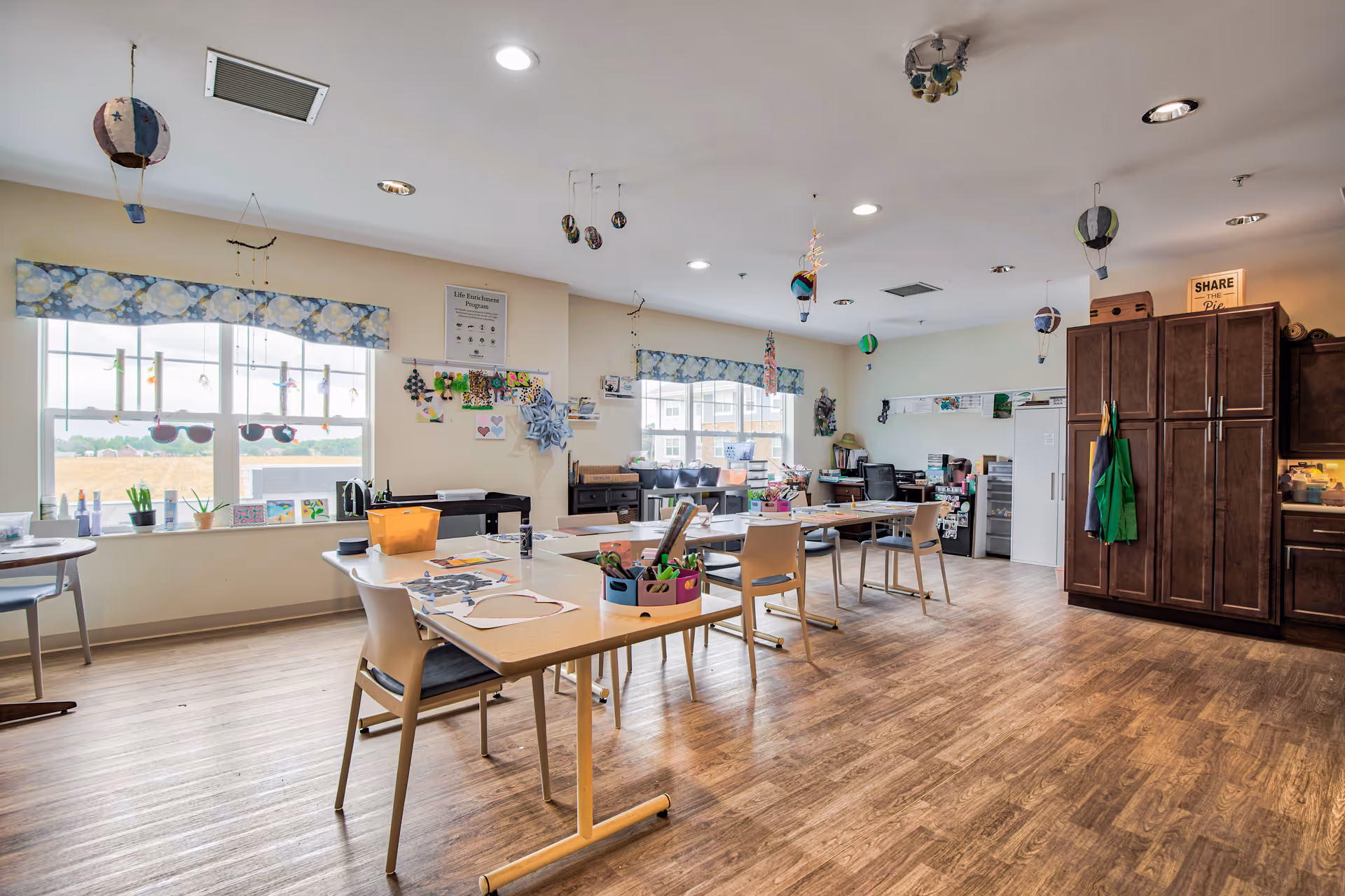 A bright, spacious activity room with wooden flooring and large windows letting in natural light. Several tables and chairs are arranged around the room with various craft supplies and decorations on the tables. Colorful hot air balloon decorations hang from the ceiling. There are cabinets and shelves along the walls, and the room has a welcoming, creative atmosphere.