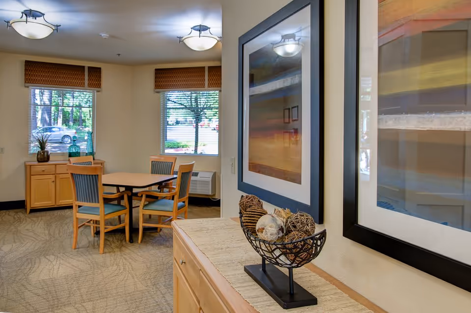 A cozy dining area in Marquis Wilsonville Assisted Living featuring a square wooden table with four chairs. Two windows with blinds and valances let in natural light, showing a view of trees and parked cars outside. A wooden sideboard with decorative vases is positioned under one window. In the foreground, a cabinet with a decorative bowl filled with woven balls and a framed abstract painting on the wall add to the warm ambiance.