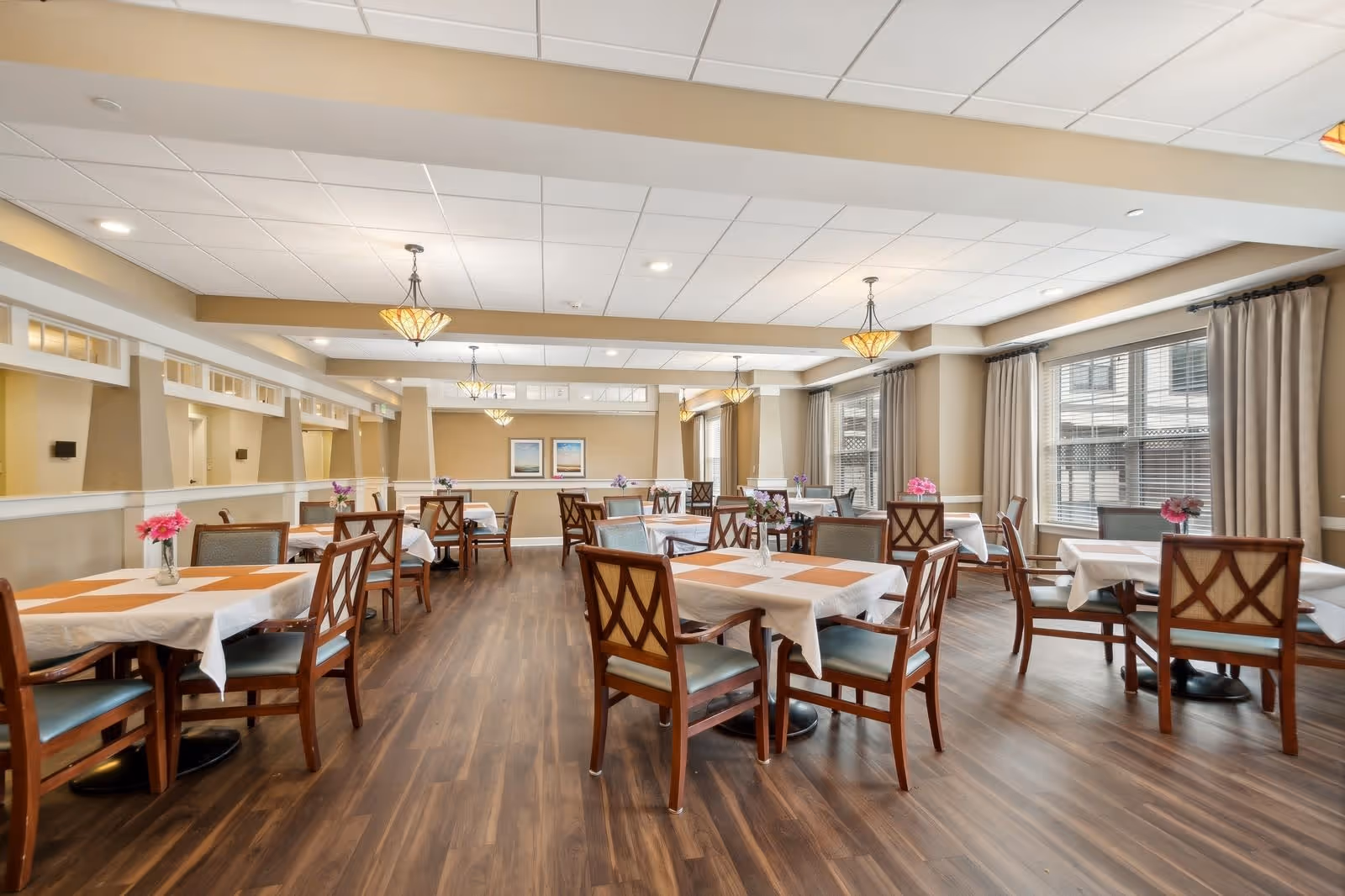A spacious dining room with multiple tables covered in white tablecloths and orange placemats. Each table is surrounded by wooden chairs with cushioned seats and backs. The room has large windows with beige curtains allowing natural light to enter. The floor is wooden, and the ceiling has recessed lighting and hanging light fixtures. There are decorative flowers on each table and framed artwork on the far wall.