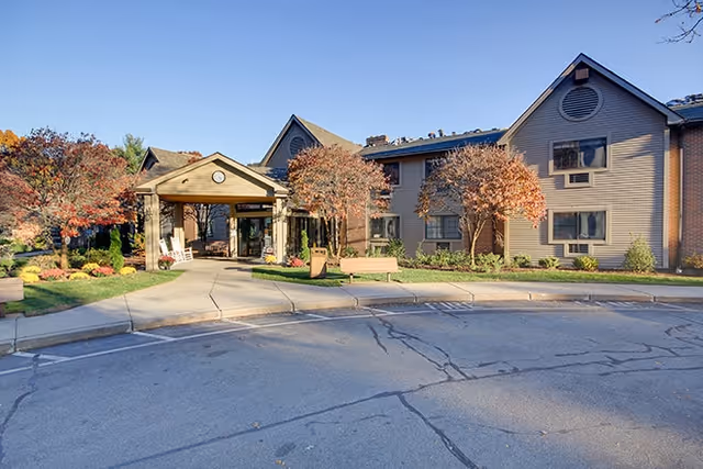 Exterior view of Smithfield Woods senior living facility showing a two-story building with a covered entrance, landscaped garden with trees and shrubs, and a paved driveway under a clear blue sky.