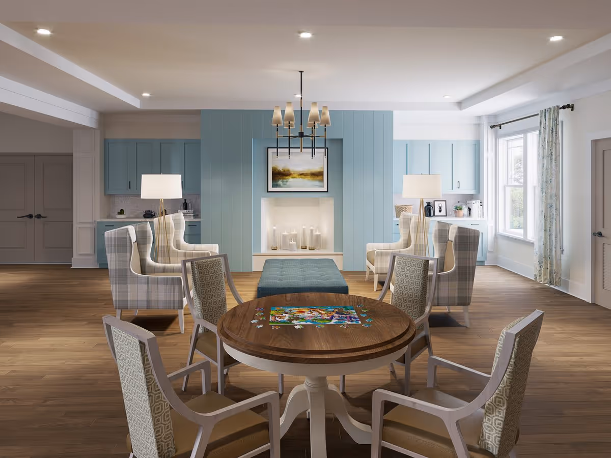 A bright and spacious common area with a round wooden table in the foreground featuring a partially completed jigsaw puzzle. Surrounding the table are four upholstered chairs with patterned fabric. In the background, there are four plaid armchairs arranged around a blue ottoman in front of a modern fireplace with candles inside. Above the fireplace hangs a landscape painting. The walls behind the fireplace are painted light blue with built-in cabinetry on either side. Two floor lamps with white shades flank the armchairs. Large windows with floral curtains allow natural light to fill the room, and the floor is covered with light wood planks.