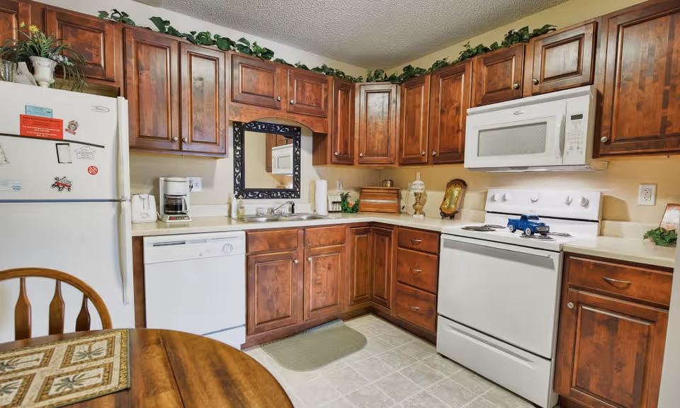 A kitchen with wooden cabinets, white appliances including a refrigerator, dishwasher, stove, and microwave. There is a coffee maker on the counter, a small blue toy truck on the stove, and decorative plants on top of the cabinets. A wooden dining table with a placemat is partially visible in the foreground.