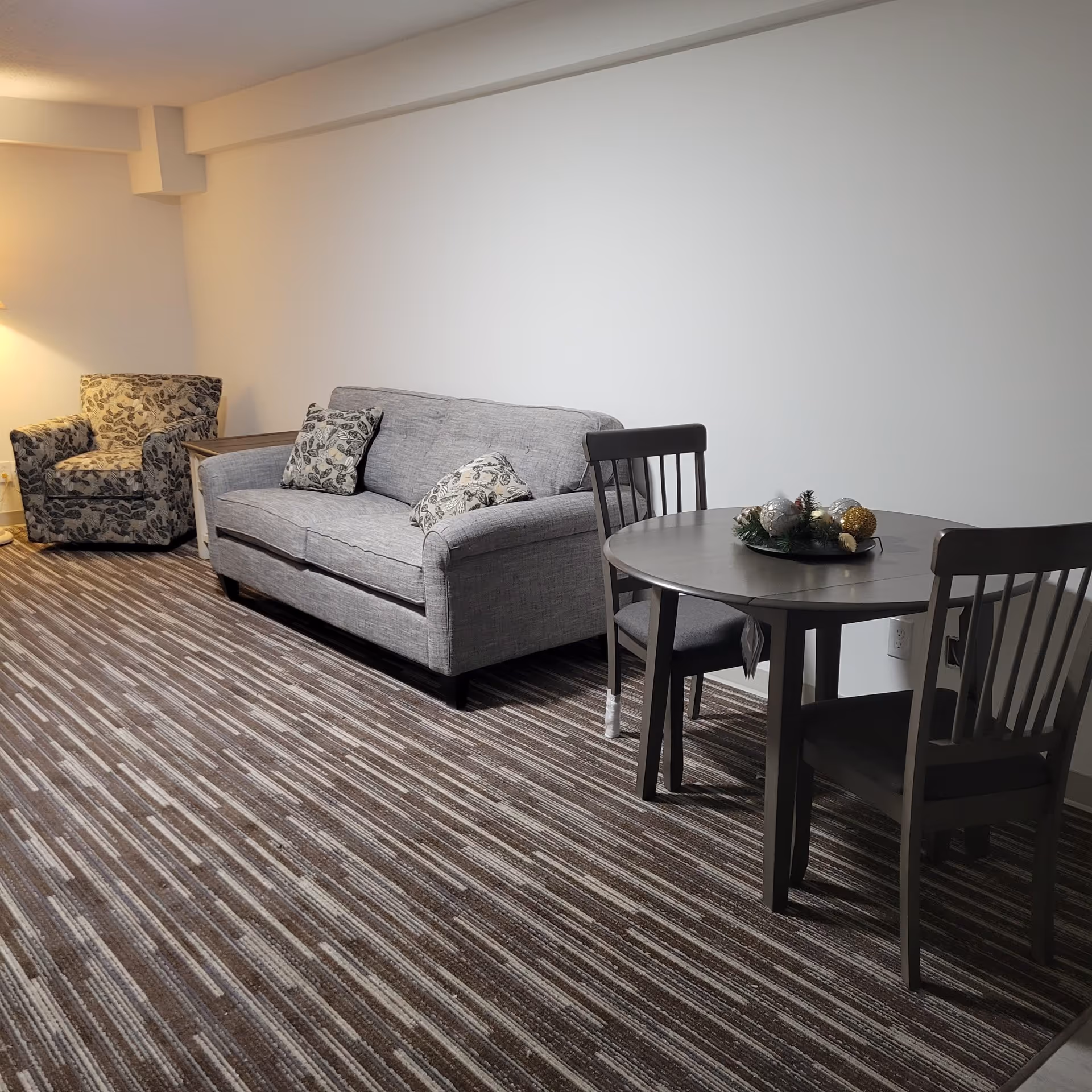 A living area with a gray sofa adorned with two patterned cushions, a matching armchair, a wooden side table, and a round dining table with two chairs. The floor is covered with a striped carpet, and there is a lamp in the corner providing warm light.