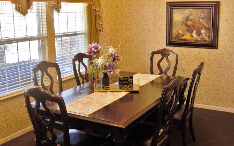 A dining room with a dark wooden table and six matching chairs. The table is decorated with a floral centerpiece and a table runner. There are three large windows with blinds and valances on the left side, and a framed painting of birds on the wallpapered wall.