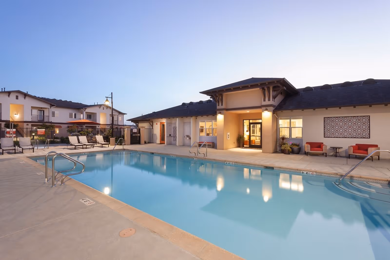 Outdoor swimming pool area at Villa del Sol senior living facility during dusk, featuring poolside lounge chairs, umbrellas, and a building with lit entrance and seating area.