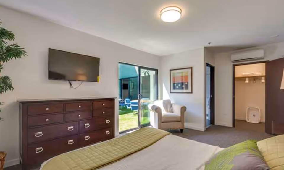 A bright bedroom with a bed covered in green and beige bedding, a dark wooden dresser with a flat-screen TV mounted above it, a beige armchair near a sliding glass door leading to an outdoor patio with blue chairs, and an open doorway showing a walk-in closet and an air conditioning unit above.