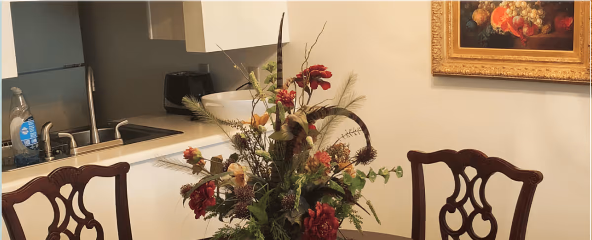 A kitchen area with a countertop featuring a decorative floral arrangement in the foreground. Behind the counter, there is a sink with a faucet, a bottle of dish soap, and a black appliance. A framed painting with fruit is hanging on the wall to the right. Two wooden chairs with ornate backs are positioned near the counter.